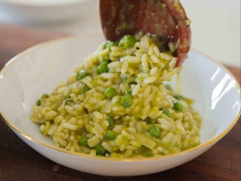 A close-up of creamy risotto with green peas being served onto a white plate with a gold rim using a wooden spoon.